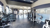 This fitness center offers a comprehensive workout environment designed to accommodate a variety of exercise needs. The space is housed on a single floor and features notably high ceilings accented by a dark upper border, creating a sense of volume and openness throughout. Large arched windows line one wall, flooding the interior with natural light and offering outside views, while ceiling fans provide additional airflow and comfort.

The flooring consists of durable, textured rubber matting in a dark tone, well-suited for heavy equipment and high-traffic use. On one side of the facility, a row of cardio machines — including treadmills and steppers — is neatly arranged to maximize floor space. The opposite side is dedicated to strength training, featuring multi-station cable machines, weight benches, and free weight equipment. Full-length mirrors are positioned along the walls to assist with form and to visually expand the space.

Recessed and flush-mounted lighting supplements the natural light, ensuring the facility remains well-lit throughout the day. The overall aesthetic is clean and functional, with a blue and white color scheme that lends a professional, gym-grade atmosphere. This fitness center is well-appointed to support both cardio and resistance training routines.