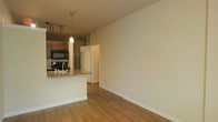 This single-floor residence offers a thoughtfully designed open-concept layout that seamlessly connects the kitchen and living areas. The kitchen is a standout feature, showcasing rich wood cabinetry, granite countertops, and a breakfast bar peninsula that provides both additional prep space and a casual dining option. Black stainless steel appliances, including a built-in microwave and range, complement the kitchen's functional design. Pendant lighting above the peninsula and a track lighting system add layered illumination, enhancing the warm ambiance of the space. The continuous hardwood-style flooring flows throughout the open area, contributing to a cohesive and inviting atmosphere. Neutral, cream-toned walls offer a clean, versatile backdrop well-suited to a variety of interior styles. A decorative column detail adds a subtle architectural touch at the kitchen's edge. The open living area provides generous room to customize the space to suit various furniture arrangements. A doorway visible from the main living area suggests additional rooms beyond, offering functional separation within the overall layout. The space strikes a balance between modern finishes and comfortable, everyday livability.