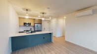 This single-floor unit showcases a thoughtfully designed open-concept layout with clean, contemporary finishes throughout. The kitchen serves as a focal point, featuring a striking teal-paneled island with a white quartz countertop and a built-in sink. Light wood cabinetry lines the walls above a geometric tile backsplash, complemented by stainless steel appliances including a gas range, over-the-range microwave, and French door refrigerator. Globe pendant lights hang above the island, adding a warm, modern touch. Light wood-toned luxury vinyl plank flooring flows seamlessly throughout the space, unifying the open living area with the kitchen.

The living area is spacious and bright, with clean white walls that create a neutral, versatile backdrop. The bedroom features a large multi-pane window offering views of surrounding greenery, flooding the room with natural light, and includes a wall-mounted mini-split HVAC unit for individual climate control. A second, smaller bedroom also benefits from a window with outdoor views.

The bathroom is well-appointed with a subway-tiled tub-shower combination, matte black fixtures, a light wood vanity with white countertop, and a frameless mirror, maintaining the unit's cohesive modern aesthetic throughout.