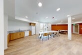 Located at 65 Shiloh Rd in Windsor, California, this single-floor community room offers a versatile and welcoming space designed for group gatherings, meetings, and events. The open-plan layout is bright and airy, with large windows that invite natural light and provide views of the surrounding neighborhood. Light wood-look flooring runs throughout, complementing the clean white walls and a bold terracotta-orange accent wall that adds warmth and visual interest to the space.

The main area is furnished with multiple mobile white tables and chairs, allowing for flexible seating arrangements to suit a variety of uses. Recessed ceiling lighting ensures the space is well-illuminated throughout the day and evening. A structural column near the center of the room adds an architectural element without disrupting the open feel.

A kitchenette along one wall is equipped with warm wood cabinetry, granite-look countertops, a stainless steel sink, a dishwasher, a full-size refrigerator, and upper storage cabinets. A dual drinking fountain and bottle-filling station is conveniently mounted near the kitchen area. The space also includes an accessible restroom finished with neutral tile walls and flooring, a wall-mounted sink, and safety grab bars, ensuring the space is welcoming to all visitors.
