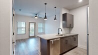 This single-floor unit showcases a contemporary design with a cohesive aesthetic carried throughout. The open-concept kitchen serves as a central feature, anchored by a spacious quartz-topped peninsula with an undermount sink and matte black faucet. Dark wood-grain cabinetry, a subway tile backsplash, and a full suite of stainless steel appliances—including a refrigerator, range, microwave, and dishwasher—complete the kitchen. Striking cone-shaped pendant lights hang above the island, adding a modern design element.

The kitchen flows seamlessly into the living area, which features wood-look plank flooring, a ceiling fan, and access to an outdoor balcony through a glass-paned door flanked by windows that allow generous natural light.

The bedroom is well-proportioned, featuring a ceiling fan and direct access to a walk-in closet and an en-suite bathroom. A second room with dual windows offers views of a landscaped outdoor area. The bathroom is finished with a wood-tone vanity, matte black fixtures, a large framed mirror, and a soaking tub surrounded by marble-style tile. Light wood-look flooring ties the spaces together throughout.