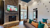 The Hawthorne Clubhouse is a thoughtfully designed community space that balances comfort and style in a welcoming, single-floor layout. The main lounge area immediately draws attention with its soaring ceilings, accented by a bold teal-painted ceiling detail and elegant crown molding. A floor-to-ceiling stacked stone fireplace serves as the room's centerpiece, topped with a wall-mounted flat-screen television — ideal for gathering and relaxing. Comfortable seating, dressed in warm teal and copper accent pillows, is arranged around a soft area rug atop hardwood flooring, creating an inviting social atmosphere. Tall French doors and transom windows allow generous natural light to pour in while offering views of the outdoor surroundings, framed by rich olive-toned draping. A separate nook area provides a more intimate setting, featuring a round dining table with banquette-style seating, decorative wall shelving with curated artwork, and a mix of modern and mid-century furnishings. Adjacent to the main lounge, a functional kitchenette includes warm wood cabinetry, granite countertops, and stainless steel appliances — well-suited for community events and everyday use. The overall ambiance is warm, modern, and welcoming throughout.