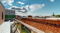 Located at 916 Main St in the heart of downtown Buffalo, New York, this rooftop terrace offers an exceptional urban outdoor experience with sweeping panoramic views of the city skyline. The terrace is thoughtfully designed to blend comfort and character, featuring a combination of large-format tile paving and wood decking that together create distinct zones for relaxation and circulation.

The main lounge area is furnished with generously sized wicker seating accented by navy blue cushions, centered around a coffee table — an inviting arrangement ideal for gatherings. A lush green hedge privacy screen lines one side of the terrace, providing a vibrant botanical backdrop that contrasts beautifully with the surrounding exposed brick architecture of neighboring historic buildings. Rustic wooden cable-rail fencing borders the perimeter, ensuring unobstructed sightlines across the Buffalo cityscape.

Raised planter beds and potted plants are integrated throughout, adding greenery and a garden-like quality to the urban setting. The classic brick exterior of the building anchors the rooftop aesthetically, reinforcing the historic character of the Main Street corridor. Under open skies, the terrace receives abundant natural light, making it a bright and dynamic outdoor retreat. An American flag display adds a patriotic touch to this well-appointed rooftop space.