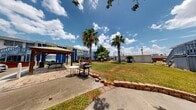 This outdoor BBQ area serves as a communal amenity space within a residential complex, designed to provide residents with a dedicated space for outdoor cooking and social gatherings. The space is thoughtfully laid out on a single level, offering a comfortable and functional environment for outdoor entertaining.

At the heart of the space is a freestanding gas grill stationed on a poured concrete pad, accompanied by a small round table for convenient prep or dining. Nearby, a wooden pergola provides a shaded retreat with additional seating and a picnic table beneath, making it an ideal spot for gatherings during sunny days. The pergola's open-roof design allows natural light to filter through while still offering a sense of shelter.

The surrounding landscape features mature shade trees, including a prominent palm tree, which contribute to a pleasant and inviting outdoor atmosphere. Brick-edged pathways and curved borders add a sense of structure and intentional design to the grounds.

The adjacent residential building is finished in a bold blue and white exterior palette with external staircases and balconies, framing the outdoor space and providing easy access for residents. A paved parking area borders the property, while green lawns and established trees complete the setting, enhancing the overall sense of community and outdoor livability.