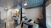 This single-story loft in Houston's 77002 zip code showcases an efficient, open-concept design with notable industrial character. The distinctive metal panel ceiling with exposed conduit runs throughout the main living area, lending an urban aesthetic that defines the space's overall ambiance.

The main living area integrates sleeping, lounging, and dining functions within a single open room. A dark curtain panel serves as a flexible room divider, subtly separating the sleeping area—which features a platform bed with a wall-mounted display shelf—from the living zone, furnished with a sofa, area rug, and a low-profile media console. A desk and chair are also positioned within the space, offering a dedicated work area. Natural light enters through a window dressed with blinds and blackout curtains.

The galley-style kitchen is clearly defined from the main living area and is well-appointed with stainless steel appliances, including a range, microwave, and refrigerator. Light wood cabinetry pairs with granite countertops and a double basin sink, providing functional workspace and ample storage.

The bathroom features light tile flooring, a wood-toned vanity with granite countertop, and clean white walls, offering a calm, neutral finish to the space.