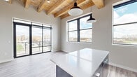 Located at 8 W 9th Ave in Oshkosh, Wisconsin, this thoughtfully designed 2-bedroom unit blends industrial character with contemporary finishes across a single floor. The open-concept living and kitchen area serves as the heart of the space, where soaring exposed wood beam ceilings and visible ductwork create a striking loft-inspired aesthetic. Large black-framed windows and a glass sliding door flood the space with natural light while offering elevated views of the surrounding Oshkosh cityscape. The kitchen is well-appointed with two-tone cabinetry — dark lower cabinets paired with crisp white uppers — stainless steel appliances including a refrigerator, range, microwave, and dishwasher, plus a sleek island with a white countertop and pendant lighting overhead. Light wood-look flooring runs throughout, complementing the neutral wall tones. The primary bedroom offers a comfortable footprint with a sliding double-panel wood closet door, while the second bedroom benefits from its own large black-framed window with neighborhood views. A hallway connects the rooms efficiently. The space combines the warmth of natural materials with clean modern design for a distinctive urban living experience.