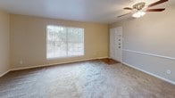 This single-story unit offers a practical and functional layout designed to maximize available space. The living area serves as the central hub of the home, finished with wall-to-wall carpet and neutral-toned walls that create a clean, versatile backdrop. A ceiling fan with a light fixture overhead provides both comfort and illumination. A large window near the front entry door allows natural light to fill the space, offering views of the surrounding greenery outside.

The kitchen is accessible via a pass-through opening with a countertop bar that connects it to the living area, making it ideal for casual dining or entertaining. White cabinetry lines the walls, complemented by granite-style laminate countertops. The kitchen is equipped with a gas range, a refrigerator, and an under-cabinet range hood, all finished in white. The flooring transitions to a hard surface in this area.

The bedroom is a quiet, carpeted space with a ceiling fan, neutral walls, and ample room for furnishings. The bathroom features a full tub and shower combination with white tile surround, a toilet, and a vanity with cabinetry. A hallway connects the various rooms, completing this straightforward and efficiently organized floor plan.