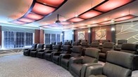 This private screening room is designed to deliver an immersive cinematic experience. The space is anchored by multiple rows of generously sized dark leather recliner chairs, each equipped with built-in cup holders, arranged in a tiered layout to ensure unobstructed sightlines for every seat. A ceiling-mounted projector serves as the primary display system, positioned centrally overhead to project onto a dedicated screen at the front of the room. The ceiling is a standout architectural feature, comprising a bold coffered design with large illuminated panels that emit warm amber and red tones, creating a dramatic, theater-like ambiance. Recessed lighting complements the colored panels, offering functional illumination throughout the space. The walls are finished in a deep, rich tone and adorned with contemporary abstract artwork, contributing to the room's sophisticated atmosphere. Patterned carpeting covers the floor, adding both comfort and a refined aesthetic. Windows along one side are fitted with slatted blinds, allowing for light control during screenings. Overall, the space combines comfort, style, and functionality into a cohesive, high-end entertainment environment.
