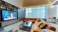 This single-floor space showcases a thoughtfully designed living area that blends contemporary aesthetics with functional elegance. The living room serves as the centerpiece, anchored by a generous sectional sofa in warm cognac leather, accented with mustard and neutral throw pillows that add depth and texture to the space.

A large flat-screen television is seamlessly integrated into a full-height built-in wall unit, which features open shelving stocked with books and decorative objects, offering both practicality and visual interest. The cabinetry is finished in a refined dark wood tone, complementing the room's overall sophisticated palette.

Floor-to-ceiling windows along one wall flood the space with natural light while providing expansive urban views of the surrounding cityscape. Sheer floor-length curtains diffuse the light softly, enhancing the room's calm and polished atmosphere. A sleek, dark rectangular coffee table grounds the seating arrangement on a subtly textured area rug.

Overhead, recessed cove lighting along the ceiling perimeter emits a warm ambient glow, adding an intimate and refined quality to the space. The overall ambiance is modern, curated, and inviting.
