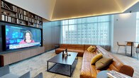 This single-floor space showcases a thoughtfully designed living area that blends contemporary aesthetics with functional elegance. The living room serves as the centerpiece, anchored by a generous sectional sofa in warm cognac leather, accented with mustard and neutral throw pillows that add depth and texture to the space.

A large flat-screen television is seamlessly integrated into a full-height built-in wall unit, which features open shelving stocked with books and decorative objects, offering both practicality and visual interest. The cabinetry is finished in a refined dark wood tone, complementing the room's overall sophisticated palette.

Floor-to-ceiling windows along one wall flood the space with natural light while providing expansive urban views of the surrounding cityscape. Sheer floor-length curtains diffuse the light softly, enhancing the room's calm and polished atmosphere. A sleek, dark rectangular coffee table grounds the seating arrangement on a subtly textured area rug.

Overhead, recessed cove lighting along the ceiling perimeter emits a warm ambient glow, adding an intimate and refined quality to the space. The overall ambiance is modern, curated, and inviting.