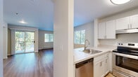This thoughtfully laid-out single-floor unit offers an efficient and functional use of space, combining a kitchen and living area into one cohesive open-plan layout. The kitchen is well-appointed with crisp white cabinetry, light countertops, a stainless steel refrigerator, an electric range with overhead ventilation, a dishwasher, and a double sink. A peninsula counter separates the kitchen from the living area, providing both additional workspace and a natural visual boundary between the two zones. The living area is bright and airy, benefiting from multiple windows fitted with blinds that invite generous natural light while offering views of the surrounding greenery. A sliding glass door opens to a private balcony, further extending the connection to the outdoors. Warm hardwood-style flooring runs throughout the main living spaces and into the bathroom, contributing to a cohesive and clean aesthetic. Built-in shelving and a closet nook offer practical storage solutions. The bathroom includes a vanity with an integrated sink, a toilet, and towel bars with brushed nickel finishes. Neutral wall tones and baseboard heating throughout give the space a comfortable, move-in-ready feel.