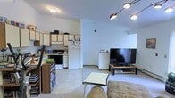 This single-floor unit features an open-concept layout that integrates the kitchen and living area into one shared space. The kitchen is equipped with white cabinetry, a black electric range, a full-size white refrigerator, and ample counter space along an L-shaped configuration. Upper cabinets provide generous storage, while the light-colored countertops contribute to a clean, functional workspace. A low dividing counter subtly separates the kitchen from the living area without fully enclosing either space.

The living area is carpeted in a neutral tone, creating a comfortable setting for everyday use. A large flat-screen television sits on a low media console, serving as the focal point of the room. A small coffee table and a cushioned sofa complete the seating arrangement, offering a relaxed atmosphere. Overhead, a multi-head track lighting fixture provides adjustable illumination, complemented by a flush-mount ceiling light in the kitchen zone.

The white walls and light flooring throughout keep the space feeling open and bright. The overall layout is straightforward and practical, making efficient use of the available footprint in a cohesive, easy-to-navigate single-level floor plan.
