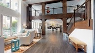 This expansive communal space makes an immediate impression with its dramatic double-height ceilings, dark hardwood flooring, and a striking framework of rich, dark timber columns and beams that anchor the open-concept layout. An interior mezzanine level with wrought-iron railings overlooks the main floor, accessible via a wood-and-iron staircase that adds architectural character throughout.

The lounge area is bathed in natural light through floor-to-ceiling glass doors and tall windows, offering views of lush outdoor greenery and palm trees. Comfortable white upholstered seating is arranged around a floral area rug, with a wall-mounted television completing the casual entertainment setup. A recessed wood-paneled ceiling accent adds warmth to this seating zone.

The kitchenette is well-appointed with white shaker cabinetry featuring glass-front upper cabinets, a dark tile backsplash, butcher block and white countertops, stainless steel appliances including a refrigerator and dishwasher, and a wine cooler. A long communal bar-height table with modern black wire stools runs alongside, illuminated by a warm chandelier.

Adjacent to the kitchen, a regulation billiards table with pendant lighting and a dark textured brick accent wall creates a dedicated game zone. Multiple dining and work tables provide flexible gathering space throughout, making this a highly functional and stylish amenity space.