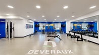 The Deveraux Fitness Suite at 3991 Spring St in Regina, Saskatchewan, is a purpose-built, single-level fitness facility offering a comprehensive workout environment within a generously sized open floor plan. The space is defined by a clean, modern aesthetic with crisp white walls accented by bold cobalt blue feature walls, creating an energetic and motivating atmosphere throughout.

Recessed pot lighting is evenly distributed across the white ceiling, ensuring bright, consistent illumination across the entire floor. The space is climate-controlled with a wall-mounted mini-split air conditioning unit, supporting year-round comfort during workouts. Natural light filters in through windows along the blue accent wall, complemented by roller blinds for adjustable brightness.

The fitness floor is well-organized and comprehensively equipped. Cardio equipment includes treadmills, an elliptical trainer, and a Concept2 RowERG rowing machine. The strength training section features a multi-function cable/cable machine, a full dumbbell rack, an adjustable weight bench, and a variety of functional training accessories including stability balls, kettlebells, and medicine balls. A large floor-to-ceiling mirror along one wall enhances the sense of space and assists users in monitoring form.

Multiple flat-screen TVs are mounted throughout, and motivational branded graphics and wall art reinforce the Deveraux identity. A water cooler station is conveniently located within the space. The Deveraux logo is prominently displayed on the floor, completing the polished, branded environment.