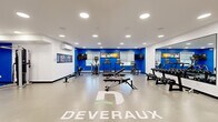 The Deveraux Fitness Suite at 3991 Spring St in Regina, Saskatchewan, is a purpose-built, single-level fitness facility offering a comprehensive workout environment within a generously sized open floor plan. The space is defined by a clean, modern aesthetic with crisp white walls accented by bold cobalt blue feature walls, creating an energetic and motivating atmosphere throughout.

Recessed pot lighting is evenly distributed across the white ceiling, ensuring bright, consistent illumination across the entire floor. The space is climate-controlled with a wall-mounted mini-split air conditioning unit, supporting year-round comfort during workouts. Natural light filters in through windows along the blue accent wall, complemented by roller blinds for adjustable brightness.

The fitness floor is well-organized and comprehensively equipped. Cardio equipment includes treadmills, an elliptical trainer, and a Concept2 RowERG rowing machine. The strength training section features a multi-function cable/cable machine, a full dumbbell rack, an adjustable weight bench, and a variety of functional training accessories including stability balls, kettlebells, and medicine balls. A large floor-to-ceiling mirror along one wall enhances the sense of space and assists users in monitoring form.

Multiple flat-screen TVs are mounted throughout, and motivational branded graphics and wall art reinforce the Deveraux identity. A water cooler station is conveniently located within the space. The Deveraux logo is prominently displayed on the floor, completing the polished, branded environment.