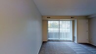This single-floor space features an open living area designed with simplicity and functionality in mind. The room is finished with neutral-toned walls and wall-to-wall carpeting in a subtle gray hue, creating a clean and understated aesthetic. A dominant feature of the space is the expansive sliding glass door spanning much of the exterior wall, dressed with full-length vertical blinds that allow for flexible light control while providing access to the outdoors. The generous width of the glazed opening allows natural light to permeate the interior, contributing to an airy atmosphere. HVAC vents are integrated into the ceiling, ensuring climate comfort throughout the space. A standard interior door is positioned along one wall, providing access to an adjacent room or storage area. The ceiling features a textured finish and maintains a consistent height across the room. The overall layout is open and unobstructed, offering flexible arrangement potential. The neutral color palette of beige walls, white trim, and gray carpet provides a versatile backdrop suitable for a variety of interior styles and furnishing choices.