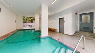 Located at 1255 Pendrell St in Vancouver's West End, this indoor pool facility offers a well-appointed aquatic space designed for both leisure and lap swimming. The pool itself is the centerpiece of the room, featuring clearly marked depth indicators along its tiled perimeter, metal entry handrails, and vivid turquoise water. The surrounding pool deck is finished in warm-toned ceramic tiles, providing safe and practical poolside footing. A wooden bench along one wall offers a convenient seating area for swimmers. The space is bright and airy, illuminated by overhead fluorescent strip lighting, while large sliding glass doors on one end flood the room with natural light and open directly onto an outdoor patio furnished with seating, framed by lush greenery. Structural columns punctuate the interior, adding architectural definition to the layout. Two grey doors provide access to changeroom or washroom facilities, and pool rules are posted on the wall for user reference. The clean white walls and tile work throughout create a crisp, functional ambiance. The overall design reflects a practical yet comfortable aquatic amenity well-suited to the needs of building residents in this vibrant Vancouver neighbourhood.