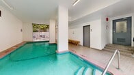 Located at 1255 Pendrell St in Vancouver's West End, this indoor pool facility offers a well-appointed aquatic space designed for both leisure and lap swimming. The pool itself is the centerpiece of the room, featuring clearly marked depth indicators along its tiled perimeter, metal entry handrails, and vivid turquoise water. The surrounding pool deck is finished in warm-toned ceramic tiles, providing safe and practical poolside footing. A wooden bench along one wall offers a convenient seating area for swimmers. The space is bright and airy, illuminated by overhead fluorescent strip lighting, while large sliding glass doors on one end flood the room with natural light and open directly onto an outdoor patio furnished with seating, framed by lush greenery. Structural columns punctuate the interior, adding architectural definition to the layout. Two grey doors provide access to changeroom or washroom facilities, and pool rules are posted on the wall for user reference. The clean white walls and tile work throughout create a crisp, functional ambiance. The overall design reflects a practical yet comfortable aquatic amenity well-suited to the needs of building residents in this vibrant Vancouver neighbourhood.
