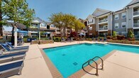 Located at 245 Creekwood Cove Ln in Lenoir City, Tennessee, this outdoor amenity area centers around a generously sized swimming pool with crystal-clear blue water, framed by a broad, well-maintained concrete sundeck. The pool area is thoughtfully designed for both relaxation and socializing, offering an ample arrangement of cushioned lounge chairs and seating grouped beneath blue market umbrellas, providing welcome shade on sunny days. A pergola structure anchors one section of the deck, creating a defined gathering spot with additional seating and a decorative fire feature nearby. The space is enclosed by a black wrought-iron fence with a keyed access gate, ensuring privacy and security for residents. Mature trees and carefully tended landscaping soften the hardscape, adding natural color and shade throughout the area. A clubhouse or amenity building with large windows is accessible directly from the pool deck, offering a seamless indoor-outdoor connection. The surrounding multi-story residential buildings feature a mix of stone accents, siding, and private balconies, creating an attractive architectural backdrop. The overall ambiance is bright, open, and resort-inspired, making this outdoor space a standout community feature within the Creekwood Cove neighborhood.