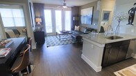 This single-floor residence offers an open-concept design that seamlessly connects the kitchen and living areas, creating a cohesive and functional layout. The kitchen features granite countertops on a peninsula island that faces the living room, providing both additional workspace and a casual bar-style seating area. Dark cabinetry adds a striking contrast to the light stone surfaces.

The living room is generously proportioned, with rich hardwood-style flooring that extends throughout the main living space, adding warmth and a polished finish underfoot. A ceiling fan with a light fixture anchors the room and promotes comfortable airflow. Tall French doors with blinds flood the space with natural light while providing direct access to a private balcony, offering views to the exterior.

Neutral gray walls are complemented by elegant floor-to-ceiling curtains in deep navy, adding depth and a refined aesthetic to the room. The space is finished with carefully chosen décor, including a bold abstract canvas and patterned area rug that tie the blue and gray palette together cohesively. A glimpse into an adjacent bedroom suggests the home continues to deliver well-appointed, comfortable living spaces throughout.