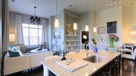 This thoughtfully designed single-floor residence offers an open-concept layout that connects the kitchen, dining, and living areas into one cohesive and inviting space. The kitchen serves as the centerpiece, featuring a large granite-topped island with an undermount sink, dark cabinetry, and a trio of cylindrical pendant lights that provide both focused task lighting and a striking visual element. The island offers ample seating, making it ideal for casual dining or entertaining.

The living area is bathed in natural light from a large window dressed with sheer white curtains, creating a bright and airy atmosphere. A decorative chandelier with black shades adds a touch of elegance to the dining nook nearby. Neutral gray walls provide a versatile backdrop throughout the space, complemented by warm hardwood-style flooring.

A built-in shelving niche along one wall offers display space for décor and personal items, adding architectural interest and functionality. The open sightlines throughout the space enhance the sense of spaciousness, while the cohesive color palette of whites, grays, and warm wood tones creates a modern yet welcoming ambiance. Overall, this residence combines contemporary design with practical living in a well-appointed, light-filled environment.