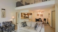 This two-story residence showcases a dramatic open-concept design anchored by soaring double-height ceilings in the main living area. An interior loft overlooks the space below via a wrought-iron railing, adding architectural interest and a sense of verticality. The main level integrates the living, dining, and kitchen areas in a seamless, flowing layout. The living room is comfortably proportioned, featuring a ceiling fan with integrated lighting and neutral-toned carpeting. A peninsula-style kitchen sits adjacent, equipped with wooden cabinetry, a microwave, refrigerator, and electric range. Pendant lighting and track lighting illuminate the kitchen workspace, while the raised breakfast bar provides an informal dining option. The dedicated dining area accommodates a full-sized table with seating for multiple guests. Warm, neutral wall tones throughout create a cohesive and inviting ambiance. The loft level, accessible via a staircase visible from the main floor, offers additional functional space overlooking the living area below, making it suitable for use as a home office, reading nook, or secondary lounge. Overall, the space balances practicality with an open, comfortable atmosphere.