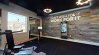 Located at Feather Sound Circle East in Clearwater, Florida, this fitness center is a thoughtfully designed, single-floor facility built to support a comprehensive workout experience. The space is divided into distinct functional zones, each catering to different training needs. The cardio area features Star Trac treadmills and other aerobic equipment positioned near large windows that allow natural light to fill the room, creating an open and energizing atmosphere. A dedicated strength and free weights zone includes a full power rack, barbells with weight plates, kettlebells, and a tiered dumbbell rack, all set on rubber flooring for safety and durability. A separate cycling and functional training area houses stationary bikes and additional cardio equipment, anchored by a striking reclaimed wood accent wall displaying the motivational phrase "Don't Wish For It, Work For It." The dark ceiling with gear-shaped ring lights adds a bold, industrial aesthetic throughout. A cable and selectorized machine section provides options for targeted muscle training. The entry area features wood-style flooring, glass doors with outdoor views, and a clean, welcoming layout. A digital fitness display is integrated into the accent wall, offering interactive workout resources. The overall design balances rugged, motivational styling with modern amenities.