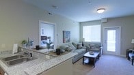 This single-floor residence offers a thoughtfully designed open-concept layout that seamlessly connects the kitchen, dining, and living areas. The kitchen features a generous granite-topped breakfast bar, providing both a practical prep surface and a casual dining space, ideal for everyday living and entertaining. The living area is comfortably sized, accommodating a full sofa and accent chairs arranged around a coffee table, and is bathed in natural light streaming through a large window fitted with horizontal blinds. Neutral-toned carpeting and soft green-gray walls throughout lend the space a calm, cohesive ambiance. A doorway visible from the main living area leads directly into a bedroom, which is furnished with a substantial bed and complementary nightstand, reinforcing the home's functional and well-organized flow. Ceiling-mounted light fixtures provide consistent ambient lighting, while HVAC vents integrated into the ceiling ensure year-round climate comfort. The overall design balances practicality with a clean, modern aesthetic, making it a comfortable and inviting single-level living space.