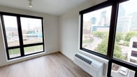 This single-floor space offers a clean, modern aesthetic defined by its bright and open atmosphere. The room features light-toned hardwood-style flooring that runs throughout, contributing to a sleek and contemporary feel. Two large, black-framed windows serve as the focal point of the space, flooding the interior with abundant natural light while framing sweeping views of an urban skyline, tree-lined streets, and surrounding city architecture. The windows provide excellent visual connectivity to the vibrant cityscape outside, making the views a standout feature of the space. The walls are finished in a neutral white tone, creating a versatile backdrop that complements a variety of design preferences. A wall-mounted HVAC unit is positioned beneath one of the windows, ensuring climate control convenience without occupying floor space. The overall layout is straightforward and functional, offering an uncluttered environment that allows the natural light and city views to take center stage. The combination of modern finishes, ample daylight, and dynamic urban vistas makes this a distinctive and well-appointed space.