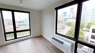 This single-floor space offers a clean, modern aesthetic defined by its bright and open atmosphere. The room features light-toned hardwood-style flooring that runs throughout, contributing to a sleek and contemporary feel. Two large, black-framed windows serve as the focal point of the space, flooding the interior with abundant natural light while framing sweeping views of an urban skyline, tree-lined streets, and surrounding city architecture. The windows provide excellent visual connectivity to the vibrant cityscape outside, making the views a standout feature of the space. The walls are finished in a neutral white tone, creating a versatile backdrop that complements a variety of design preferences. A wall-mounted HVAC unit is positioned beneath one of the windows, ensuring climate control convenience without occupying floor space. The overall layout is straightforward and functional, offering an uncluttered environment that allows the natural light and city views to take center stage. The combination of modern finishes, ample daylight, and dynamic urban vistas makes this a distinctive and well-appointed space.