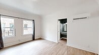 This thoughtfully designed single-floor unit makes efficient use of its intimate footprint, combining a living room, kitchen, bathroom, and hallway into a cohesive and functional layout. Light-toned hardwood flooring flows throughout the main living area, complementing the crisp white walls and clean architectural lines that give the space a bright, airy feel. A wall-mounted mini-split unit provides climate control in the living room, which also benefits from natural light streaming through a window framed by dark curtains. The kitchen is a standout feature, showcasing sage green shaker-style cabinetry with matte black hardware, white subway tile backsplash, and light quartz countertops. Stainless steel appliances — including a gas range, over-the-range microwave, and refrigerator — are paired with a deep single-basin sink finished with a sleek black faucet. An ornate black chandelier adds a touch of character above the kitchen area. The hallway connects to a dedicated vanity nook finished in a deep teal-green, complete with a mirror and built-in cabinetry, which leads into the bathroom. The bathroom features white penny tile flooring, a full tub-shower combination, and a bright window that enhances the clean, minimal aesthetic throughout.