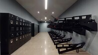This single-floor storage and equipment room is purpose-built to house a fleet of rowing machines and associated hardware in an organized, efficient layout. The elongated rectangular space features concrete masonry unit walls painted in a cool grey tone, polished concrete flooring, and a ceiling lined with recessed and linear LED lighting that ensures consistent, even illumination throughout.

Along one wall, a substantial bank of black metal lockers provides secure personal storage for users. Opposite the lockers, a series of wall-mounted rowing machine bodies are affixed in a uniform row, each unit featuring distinctive black frames and red-accented handles. Corresponding seat rail assemblies are positioned along the floor directly in front of the wall mounts, maintaining an organized, ready-to-use configuration.

The walls also feature exposed conduit runs with mounted electrical junction boxes and outlets, reflecting a practical, industrial approach to the infrastructure. The open floor plan between the equipment rows allows for easy movement and access throughout the room. The overall design prioritizes functionality and durability, with materials and finishes selected to withstand heavy, repeated use in a high-activity environment.