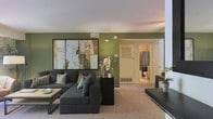 This single-floor space centers around a well-appointed living area that combines comfort with contemporary style. The room is distinguished by its bold olive-green accent walls, which create a warm and grounded ambiance. A large, dark sectional sofa serves as the focal point of the living area, complemented by coordinating accent pillows and a low-profile coffee table with a light wood surface. An oversized abstract artwork in earth tones and blues anchors the main wall, adding a refined artistic touch to the space. The carpeted flooring in a neutral beige tone adds warmth underfoot and ties the room's color palette together. A dark console table positioned along one wall provides additional surface space, paired with a small potted plant for a touch of greenery. Ceiling-mounted HVAC vents and a baseboard heating unit indicate climate control throughout. A hallway visible from the living area leads directly to a bathroom, which features a shower with a dark curtain and tiled surroundings. The layout is efficient and open, offering a functional and cohesive living environment.