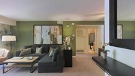 This single-floor space centers around a well-appointed living area that combines comfort with contemporary style. The room is distinguished by its bold olive-green accent walls, which create a warm and grounded ambiance. A large, dark sectional sofa serves as the focal point of the living area, complemented by coordinating accent pillows and a low-profile coffee table with a light wood surface. An oversized abstract artwork in earth tones and blues anchors the main wall, adding a refined artistic touch to the space. The carpeted flooring in a neutral beige tone adds warmth underfoot and ties the room's color palette together. A dark console table positioned along one wall provides additional surface space, paired with a small potted plant for a touch of greenery. Ceiling-mounted HVAC vents and a baseboard heating unit indicate climate control throughout. A hallway visible from the living area leads directly to a bathroom, which features a shower with a dark curtain and tiled surroundings. The layout is efficient and open, offering a functional and cohesive living environment.