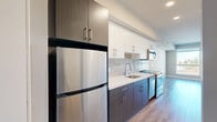 Located at 215 W 14th St in Los Angeles, CA, this efficiently designed studio offers a thoughtful layout across a single floor, making excellent use of every square foot. The galley-style kitchen is a standout feature, showcasing two-tone cabinetry with dark lower cabinets and crisp white upper cabinets, a light subway tile backsplash, quartz countertops, and a full suite of stainless steel appliances including a refrigerator, electric range, built-in microwave, and dishwasher. A dedicated laundry closet with in-unit washer is conveniently tucked just off the kitchen. The living area is bathed in natural light thanks to large, floor-to-ceiling windows that frame views of the surrounding Los Angeles cityscape. Light-toned wood-style flooring runs throughout, lending warmth and continuity to the space. The bathroom features a clean, minimalist design with a tub-shower combination, wood-look tile flooring, and bright white finishes. Two closets provide practical storage solutions. The overall aesthetic is modern and neutral, with warm beige walls, recessed lighting, and clean architectural lines that create a functional and inviting urban living space.