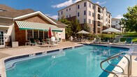 This outdoor amenity space centers around a large, well-maintained swimming pool with clearly marked depth indicators, offering both shallow and deeper sections to accommodate a range of swimmers. The pool deck is finished in a smooth concrete surface and provides substantial space for lounging, furnished with numerous green-and-white chaise lounges, chairs, and tables shaded by striped market umbrellas. A dedicated hot tub is positioned at one end of the pool, accessible via stainless steel railings.

Adjacent to the pool, a single-story cabana structure with stone veneer accents and a distinctive green-and-white striped retractable awning serves as a shaded retreat and functional support facility. Large glass-panel doors open outward, enhancing indoor-outdoor connectivity. The entire pool area is enclosed by a black iron safety fence, providing security while preserving sightlines to the surrounding mature tree canopy, which offers a natural, park-like ambiance.

The backdrop includes a multi-story residential building constructed with a combination of stucco and stone facades, adding an architectural presence to the setting. The overall environment is bright and open, benefiting from abundant natural sunlight and a well-landscaped perimeter that enhances the resort-style character of the space.