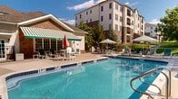 This outdoor amenity space centers around a large, well-maintained swimming pool with clearly marked depth indicators, offering both shallow and deeper sections to accommodate a range of swimmers. The pool deck is finished in a smooth concrete surface and provides substantial space for lounging, furnished with numerous green-and-white chaise lounges, chairs, and tables shaded by striped market umbrellas. A dedicated hot tub is positioned at one end of the pool, accessible via stainless steel railings.

Adjacent to the pool, a single-story cabana structure with stone veneer accents and a distinctive green-and-white striped retractable awning serves as a shaded retreat and functional support facility. Large glass-panel doors open outward, enhancing indoor-outdoor connectivity. The entire pool area is enclosed by a black iron safety fence, providing security while preserving sightlines to the surrounding mature tree canopy, which offers a natural, park-like ambiance.

The backdrop includes a multi-story residential building constructed with a combination of stucco and stone facades, adding an architectural presence to the setting. The overall environment is bright and open, benefiting from abundant natural sunlight and a well-landscaped perimeter that enhances the resort-style character of the space.