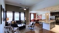 This two-story space showcases an open-concept layout that seamlessly connects the living, dining, and kitchen areas into one cohesive, flowing environment. Rich hardwood flooring extends throughout the main level, anchoring the space with warmth and continuity. The living area features elevated ceilings fitted with a ceiling fan, and large windows dressed with dark curtain panels that allow generous amounts of natural light to filter in while adding a dramatic contrast to the walls.

A bold, deep brown accent wall defines the living area, which is furnished with a dark leather sofa and patterned accent chairs, creating a striking visual dynamic. Adjacent to the living space, a dining area is highlighted by a vibrant red-toned table setting and complementary wall art, adding a pop of color to the interior. Track lighting overhead further illuminates this zone.

The kitchen bar area transitions naturally from the dining space, featuring granite countertops and pendant lighting hanging above bar-height seating. The overall ambiance of the main level is sophisticated and inviting, balancing bold color choices with open, airy proportions that give the space a comfortable yet stylish character.