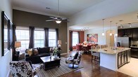 This two-story space showcases an open-concept layout that seamlessly connects the living, dining, and kitchen areas into one cohesive, flowing environment. Rich hardwood flooring extends throughout the main level, anchoring the space with warmth and continuity. The living area features elevated ceilings fitted with a ceiling fan, and large windows dressed with dark curtain panels that allow generous amounts of natural light to filter in while adding a dramatic contrast to the walls.

A bold, deep brown accent wall defines the living area, which is furnished with a dark leather sofa and patterned accent chairs, creating a striking visual dynamic. Adjacent to the living space, a dining area is highlighted by a vibrant red-toned table setting and complementary wall art, adding a pop of color to the interior. Track lighting overhead further illuminates this zone.

The kitchen bar area transitions naturally from the dining space, featuring granite countertops and pendant lighting hanging above bar-height seating. The overall ambiance of the main level is sophisticated and inviting, balancing bold color choices with open, airy proportions that give the space a comfortable yet stylish character.