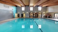 This single-story facility is defined by its striking architectural framework, most notably the warm, exposed wood beam ceilings that span the length of the structure, lending a natural, lodge-like character throughout. The centerpiece of the space is a generously sized indoor swimming pool with clear turquoise water, surrounded by tile flooring and framed by tall clerestory windows that draw in abundant natural light. The pool area features smooth concrete walls rising to meet the wood-planked ceiling, creating an open, airy atmosphere with a sense of considerable volume and height.

Adjacent to the pool, lounge areas provide casual seating with contemporary outdoor-style sectional sofas and daybeds accented with striped cushions. These relaxed gathering spaces are finished with white paneled walls and tile flooring, and feature large sliding glass doors that open directly onto an exterior patio furnished with umbrella-shaded tables and chairs, offering views of the surrounding outdoor grounds. The interior and exterior connect seamlessly, encouraging indoor-outdoor use.

A long corridor with white paneled walls, wall-mounted sconce lighting, and baseboard heating runs along one side of the facility, providing functional circulation between areas. Overall, the space balances recreational utility with a warm, inviting ambiance.