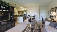This single-floor residence offers an open-concept layout that effortlessly connects the main living, dining, and kitchen areas into one cohesive and functional space. The kitchen is equipped with stainless steel appliances, including a refrigerator, microwave, and range, and features dark cabinetry paired with a dark stone countertop peninsula that doubles as a breakfast bar with seating. Recessed and overhead lighting illuminates the workspace, while a hanging pendant light adds a decorative touch above the dining area. The dining space comfortably accommodates a dark wood table and chair set, positioned beneath a classic pendant fixture. The living area flows naturally from the dining space, finished with neutral-toned carpet that adds warmth underfoot and is complemented by light-colored walls throughout. A dark wood entertainment unit anchors one side of the living room, adding a sense of elegance. The layout is practical and well-proportioned, with clear sightlines across each zone, making the space feel open and airy. An entry door with white paneling is visible, contributing to the clean, classic aesthetic. Overall, the residence presents a comfortable and stylish interior suited for everyday living.