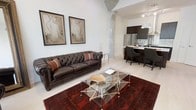This single-floor space showcases a compelling blend of industrial and contemporary design. The open-concept layout seamlessly connects the living area and kitchen, creating an expansive, airy feel. Exposed concrete columns add a distinctive industrial character, complementing the crisp white walls and light flooring throughout.

The living area is anchored by a rich, dark brown tufted leather Chesterfield sofa, paired with a striking geometric glass and chrome coffee table resting atop a deep red patterned area rug. A pair of framed figurative artwork pieces adorn the walls, adding an artistic touch to the refined atmosphere.

The kitchen is finished with dark cabinetry and stainless steel appliances, including a full-size refrigerator. A kitchen island with seating for multiple guests serves as both a functional workspace and casual dining spot, with pendant lighting overhead providing warm, focused illumination. Track lighting along the ceiling ensures the entire space is well-lit.

Overall, the space exudes a sophisticated urban ambiance, combining raw architectural elements with polished furnishings. The open flow between living and kitchen areas makes it well-suited for both everyday living and entertaining.