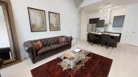 This single-floor space showcases a compelling blend of industrial and contemporary design. The open-concept layout seamlessly connects the living area and kitchen, creating an expansive, airy feel. Exposed concrete columns add a distinctive industrial character, complementing the crisp white walls and light flooring throughout.

The living area is anchored by a rich, dark brown tufted leather Chesterfield sofa, paired with a striking geometric glass and chrome coffee table resting atop a deep red patterned area rug. A pair of framed figurative artwork pieces adorn the walls, adding an artistic touch to the refined atmosphere.

The kitchen is finished with dark cabinetry and stainless steel appliances, including a full-size refrigerator. A kitchen island with seating for multiple guests serves as both a functional workspace and casual dining spot, with pendant lighting overhead providing warm, focused illumination. Track lighting along the ceiling ensures the entire space is well-lit.

Overall, the space exudes a sophisticated urban ambiance, combining raw architectural elements with polished furnishings. The open flow between living and kitchen areas makes it well-suited for both everyday living and entertaining.
