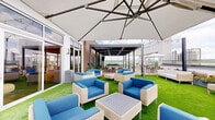 Located at 100 Colonial Homes Dr in Atlanta's vibrant Buckhead neighborhood, this rooftop terrace offers a thoughtfully designed outdoor amenity space with stunning panoramic views of the Atlanta skyline. The terrace is divided into several distinct zones, each catering to a different style of outdoor living and socializing. A lush artificial turf lawn anchors the primary lounge area, furnished with wicker seating and bold blue cushions arranged beneath large cantilevered umbrellas, creating a comfortable and inviting atmosphere. Adjacent to this area, a sleek modern pergola with louvered panels provides a shaded retreat with additional lounge seating, string lights overhead, and open sightlines to the cityscape beyond. A dedicated sun deck features wood-tile flooring and patio dining sets shaded by freestanding umbrellas, ideal for alfresco meals with skyline backdrops. Planters with colorful florals add natural accents throughout the space. Glass panels along the perimeter preserve unobstructed views while providing wind protection. The terrace connects seamlessly to an interior space through large glass sliding doors, blending indoor and outdoor living. The overall ambiance is modern, polished, and resort-inspired, making it a premier outdoor gathering destination.