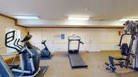 This fitness center in Birmingham, Alabama offers a well-organized, single-room exercise space designed to accommodate a variety of workout routines. The room features neutral-toned walls accented by a warm wood crown molding border that runs along the perimeter, contributing to a clean and polished aesthetic. Durable carpet flooring covers the entire space, providing cushioning underfoot during workouts.

The facility is equipped with a comprehensive selection of exercise equipment. Cardio machines include multiple treadmills — among them a Vision Fitness model — an elliptical trainer, and a stationary bike, all positioned on protective floor mats. A Hoist multi-station cable strength system anchors the strength training area, offering users options for a range of resistance exercises.

Overhead fluorescent lighting ensures the space is consistently and evenly lit throughout. A glass-paneled exterior door on one wall allows natural light to filter into the room and provides a view to the outdoors, adding a sense of openness. Additional interior doors offer connectivity to the surrounding facility. Informational signage, including exercise room rules, is posted on the walls. A fire extinguisher is also present, reflecting attention to safety. Overall, the fitness center delivers a functional and accessible environment for daily exercise.
