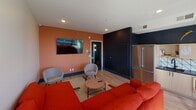 This single-floor space makes a striking first impression with its bold, contemporary design and carefully curated color palette. The open-plan layout seamlessly connects the living area and kitchen, creating a cohesive and inviting environment throughout.

The living area is anchored by a vibrant terracotta-orange accent wall, which pairs dynamically with deep navy-blue architectural elements. Recessed lighting illuminates the space from above, complementing the light wood-look flooring that runs throughout. A wall-mounted flat-screen television and comfortable seating arrangement make this an ideal space for gathering and relaxation.

The kitchen is a standout feature, showcasing light wood cabinetry with matte black hardware set against a dramatic dark navy upper wall. A geometric patterned tile backsplash with angular, graphic lines adds visual interest, while the dark countertops provide a sophisticated contrast. A stainless steel French door refrigerator and built-in appliances complete the well-appointed culinary space. A kitchen island with bar seating serves as both a functional workspace and a natural gathering point.

Floor-to-ceiling windows and a glass door open to a private balcony, flooding the interior with abundant natural light and offering open panoramic views of the surrounding cityscape, enhancing the overall sense of openness and connection to the outdoors.