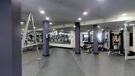 This fitness center offers a comprehensive workout environment designed to accommodate a wide range of exercise routines. The space is anchored by a series of bold purple structural columns that add a distinctive visual character to the room. Large floor-to-ceiling mirrors line the walls, creating a sense of depth and allowing users to monitor their form throughout their workouts. The flooring consists of durable rubber gym matting, providing both traction and cushioning underfoot. A full row of cardio equipment, including treadmills and elliptical machines, is arranged along one wall, with mounted flat-screen televisions positioned for easy viewing during use. The free weights section features a well-organized dumbbell rack stocked with a variety of weights, accompanied by an adjustable bench for targeted strength training. Recessed ceiling lighting ensures the entire space is evenly and brightly illuminated. The open layout promotes ease of movement between stations, making the facility functional for both individual and simultaneous group use. Overall, the fitness center presents a clean, professional atmosphere well-suited to a broad range of fitness goals and experience levels.