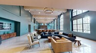 Located on Elm Street in Hartford, CT, this expansive single-floor venue blends historic architectural character with sophisticated modern design. The space is defined by soaring double-height ceilings, dramatic arched windows with ornate detailing, and a palette of muted teal and gray walls that create a refined, welcoming ambiance. Polished terrazzo flooring extends throughout, reflecting natural light that pours in generously through the oversized windows, offering views of the surrounding greenery.

The main gathering area is thoughtfully appointed with a large sectional seating arrangement, lounge chairs, and a central coffee table, all anchored by a statement area rug. A substantial communal dining table with upholstered chairs provides a formal yet inviting focal point, complemented by a bar counter with stylish stools along one wall. Overhead, circular chandelier fixtures cast warm, layered lighting across the open floor plan.

A grand staircase with rich wood cladding and sleek glass railings serves as a visual centerpiece, leading to an upper-level mezzanine with glass balustrades that overlook the main floor. Arched alcoves add architectural depth, housing media displays and decorative furnishings. The combination of historic detailing—evident in the patterned window glass and arched forms—with contemporary finishes and furnishings gives this space a distinctive and versatile character.