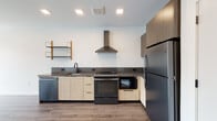 Located at 144 Addison Street in Boston's East Boston neighborhood, this single-floor unit showcases a clean, modern aesthetic throughout. The open layout is anchored by light wood-look flooring that runs continuously across the living and kitchen areas, contributing to a cohesive and airy feel.

The kitchen is thoughtfully designed with two-tone cabinetry, pairing light wood lower cabinets with darker upper cabinets and a matching dark countertop. A stainless steel range hood serves as a focal point above the electric range, complemented by a full suite of appliances including a stainless refrigerator, dishwasher, and built-in microwave. Open wood shelving with metal brackets adds a functional yet stylish accent to the kitchen wall.

The living area benefits from recessed lighting and a large window that draws in natural light while offering views of the surrounding neighborhood. The clean white walls and minimal detailing enhance the sense of openness within the space.

The bathroom features warm-toned square tile flooring, a dark wood floating vanity with an integrated sink and illuminated mirror, black matte hardware and towel bars, and a glass-enclosed shower, creating a refined and modern atmosphere throughout.