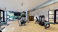 This single-level fitness center offers a thoughtfully designed workout environment that balances functionality with modern aesthetics. The space is organized to accommodate a wide variety of exercise needs, with distinct zones dedicated to cardiovascular training and strength work.

The cardio area features a row of treadmills and elliptical machines positioned along a windowed wall, allowing natural light to fill the space and providing views to the outdoors. Full-length mirrors are strategically placed to enhance the sense of openness and assist users during exercise. A dedicated free weights section includes a complete dumbbell rack and flat bench, finished with dark rubber flooring matting for safety and durability.

The strength training zone houses multiple selectorized cable and resistance machines with green upholstered padding, offering a cohesive visual identity. Warm wood-toned open shelving units serve as functional dividers throughout the space. Light wood-effect flooring runs throughout, contributing to a clean and contemporary feel.

Overhead, exposed metallic ventilation ducts run along the white ceiling alongside cylindrical pendant lighting fixtures, lending an industrial character to the design. A bold geometric green-toned mural decorates one accent wall, adding visual energy. The overall ambiance is bright, open, and purpose-driven.